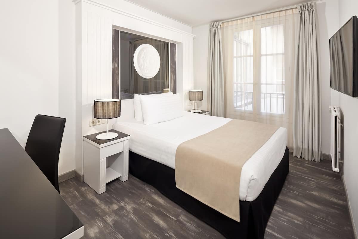 A hotel room with a large bed, two bedside tables with lamps, a round mirror, a window with curtains, and a view of the outdoors.