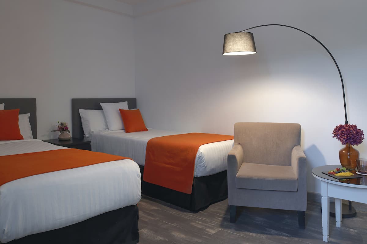 A hotel room with two beds, a chair, a table with a vase and a plate of food, and a lamp.