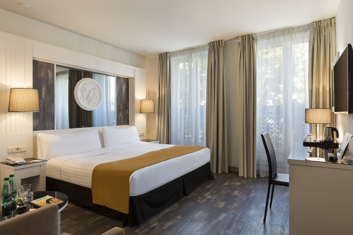 A hotel room with a large bed, bedside lamps, a desk with a chair, and a view of the outdoors through sheer curtains.