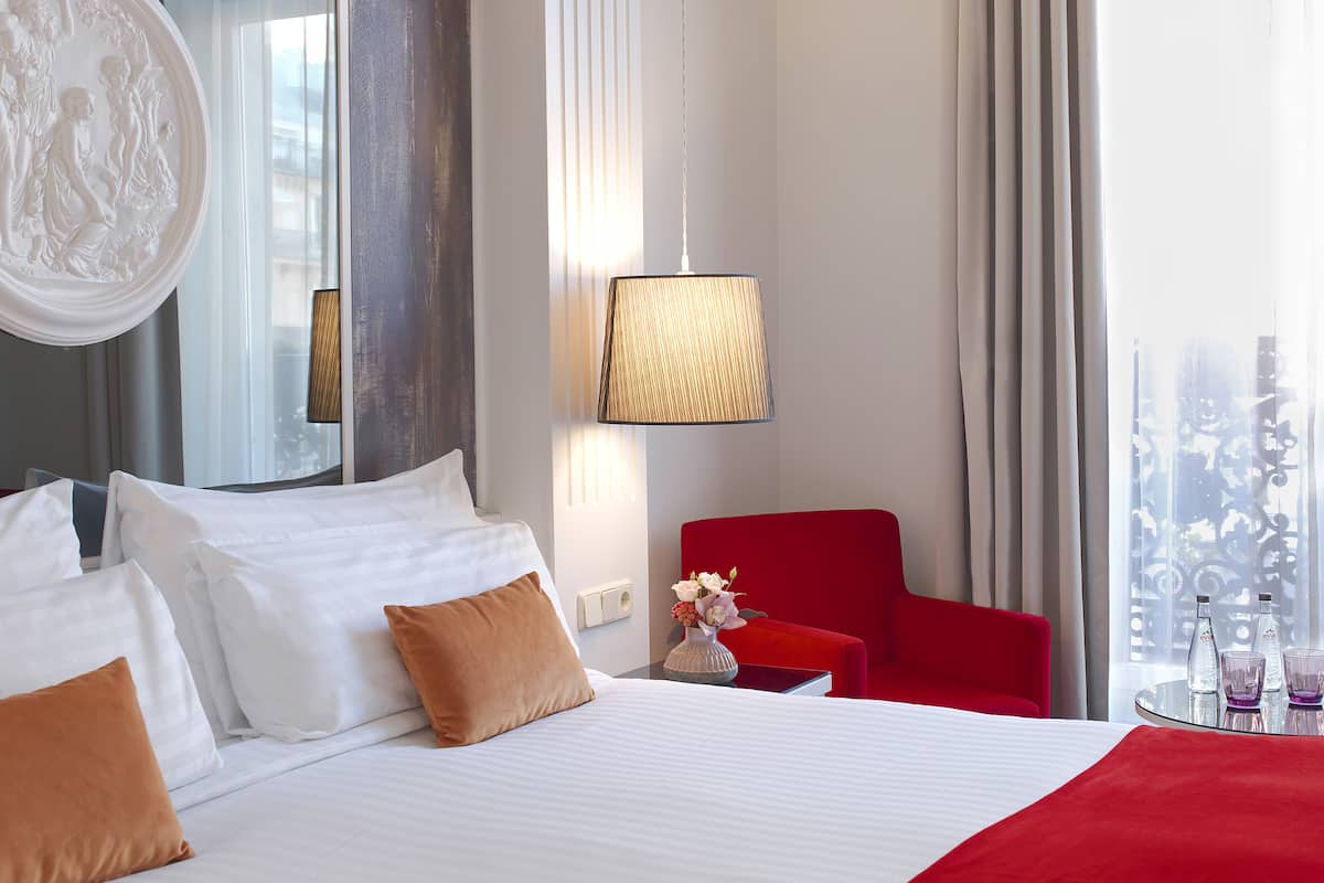 A hotel room with a large bed, a red armchair, a table with glasses and bottles, and a window with a view.