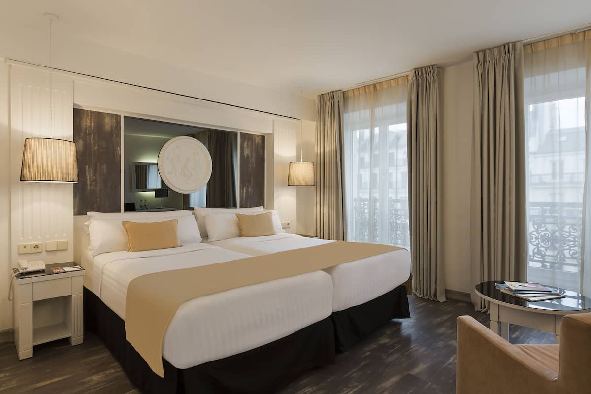 A hotel room with a large bed, bedside tables, a desk, a chair, and a view of the city through large windows.