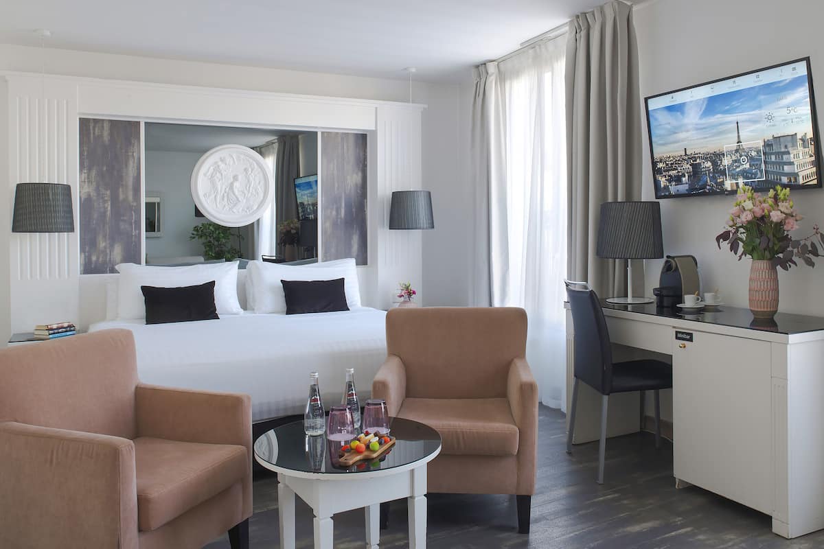 A hotel room with a bed, two armchairs, a desk with a chair, a TV, and a table with bottles and glasses.