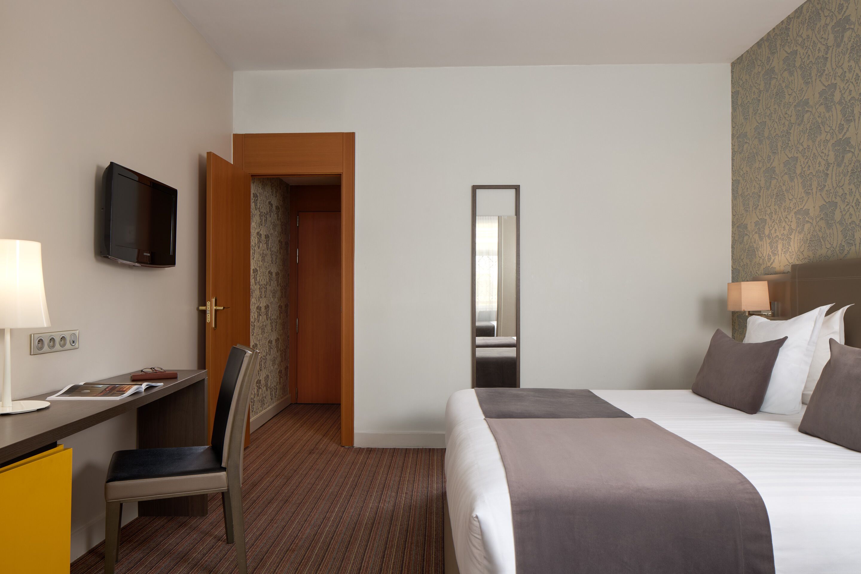 Una habitación de hotel con cama, escritorio, silla, televisor y espejo.