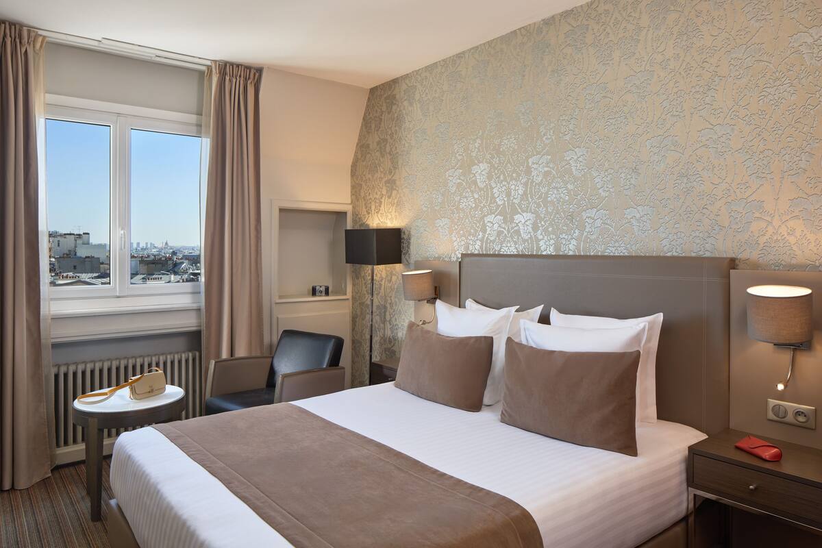 Habitación de hotel con una cama grande, un escritorio, una silla y vistas a la ciudad a través de una ventana.