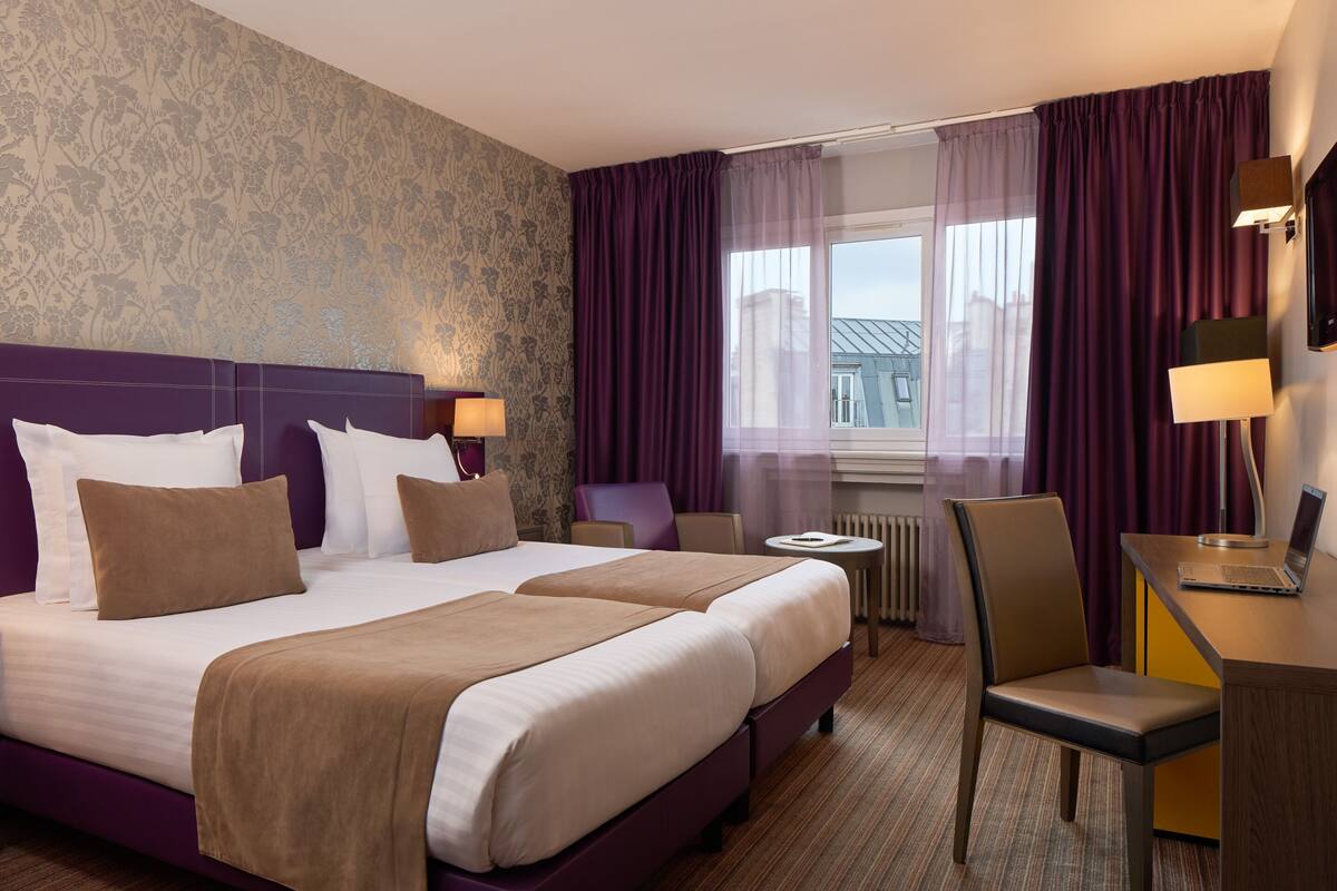 Habitación de hotel con una cama grande, dos mesitas de noche, un escritorio con una computadora portátil, una silla y vistas a la ciudad a través de la ventana.