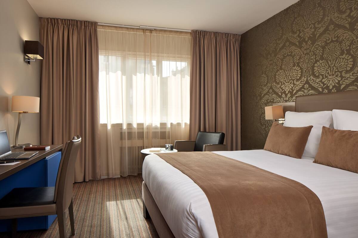 Habitación de hotel con una cama grande, un escritorio con una computadora portátil, una silla, una mesita y una lámpara.