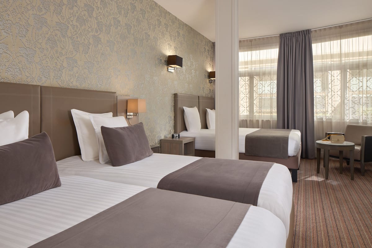 Habitación de hotel con dos camas, una mesita, una silla y vistas al exterior a través de cortinas traslúcidas.