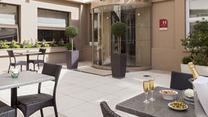 A hotel exterior with a glass entrance, outdoor seating area, and a champagne bottle on a table. - Timhotel Opera Blanche Fontaine (Paris)