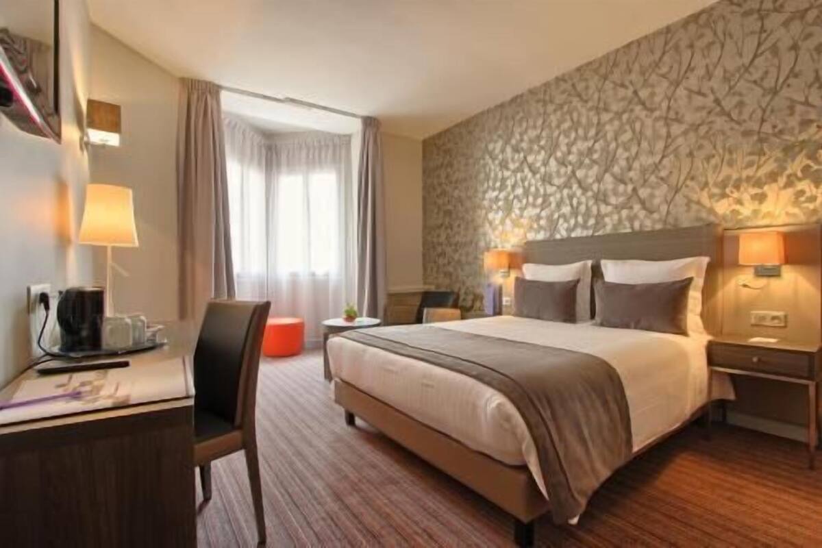 Habitación de hotel con cama, dos mesitas de noche con lámparas, un escritorio con lámpara, una silla y una ventana con cortinas.