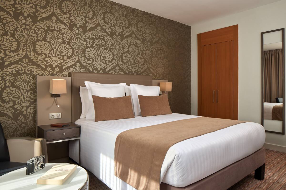 Habitación de hotel con una cama grande, dos lámparas de noche, una mesita de noche, una mesa redonda con un libro y una cámara, y un armario.