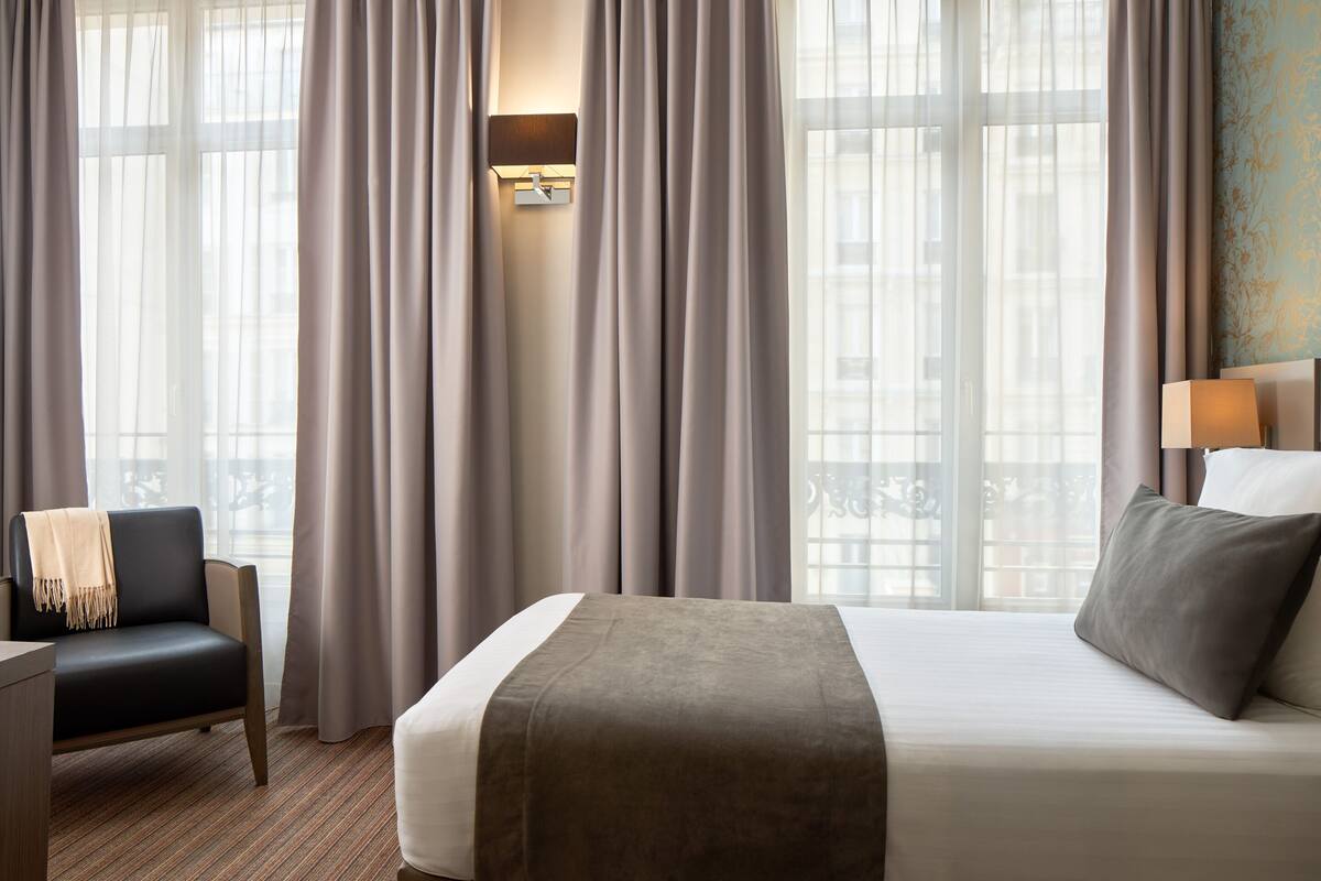 Habitación de hotel con cama, una silla, escritorio y ventana con cortinas.