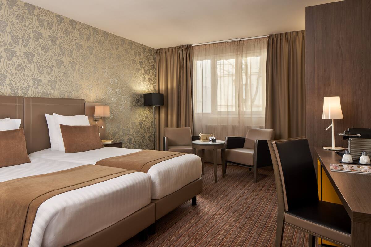 Habitación de hotel con dos camas, un escritorio, una silla, una lámpara y una ventana con cortinas.