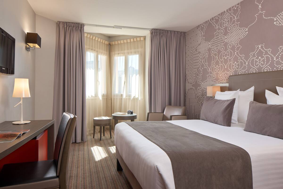 Habitación de hotel con una cama grande, un escritorio con lámpara, una silla, una mesita y un televisor montado en la pared.