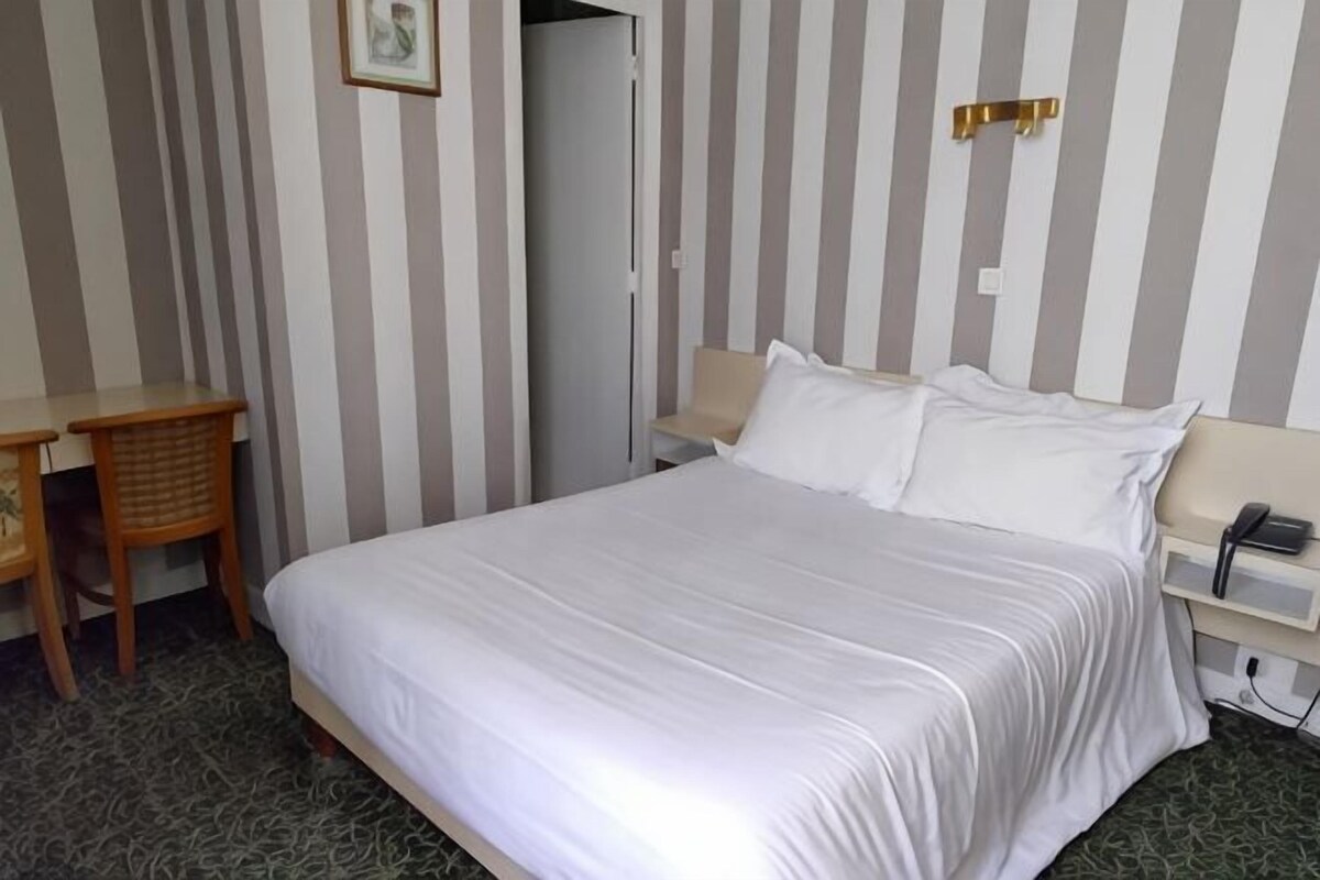 A hotel room with a bed, a desk, a chair, a bedside table, and a phone.