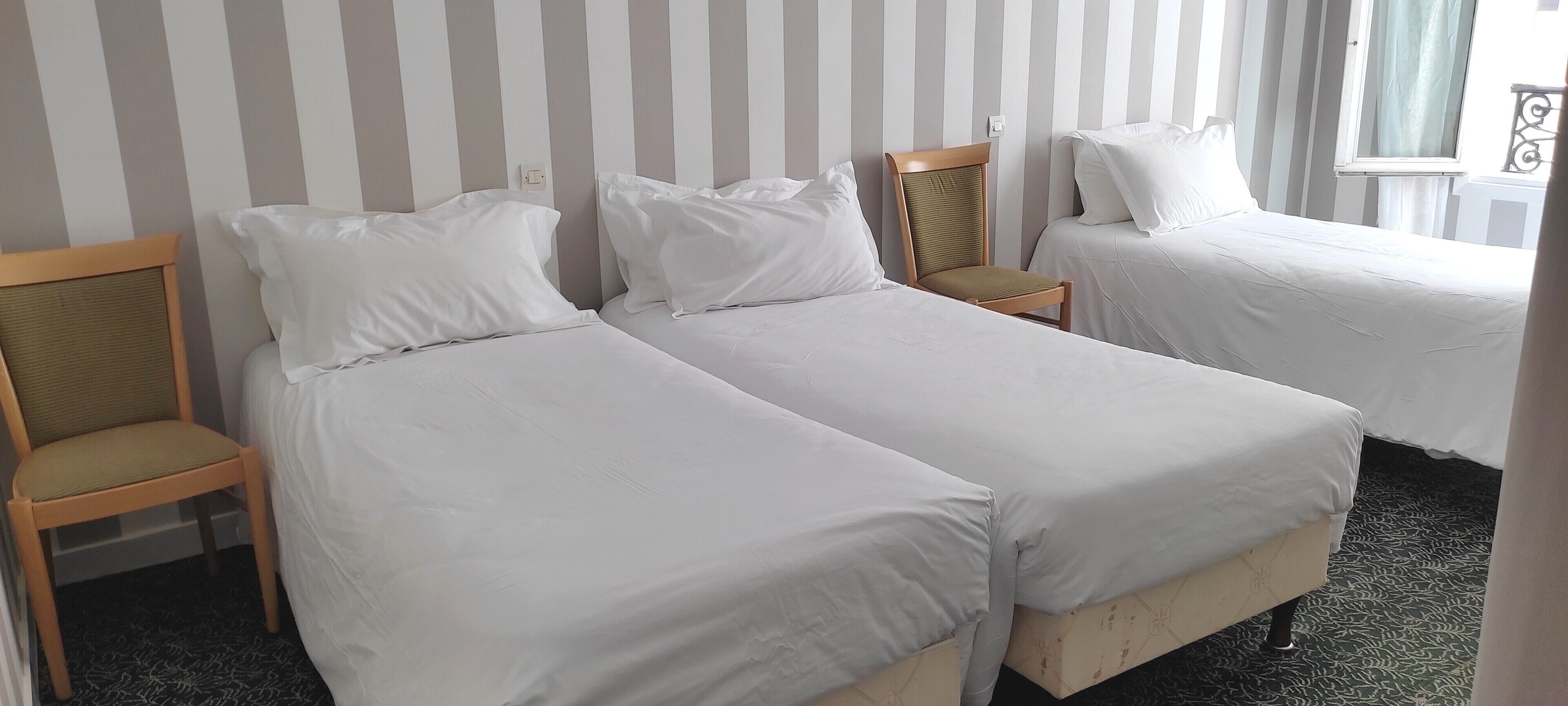 Quarto de hotel com duas camas de solteiro, uma cadeira de madeira e uma janela com cortinas.