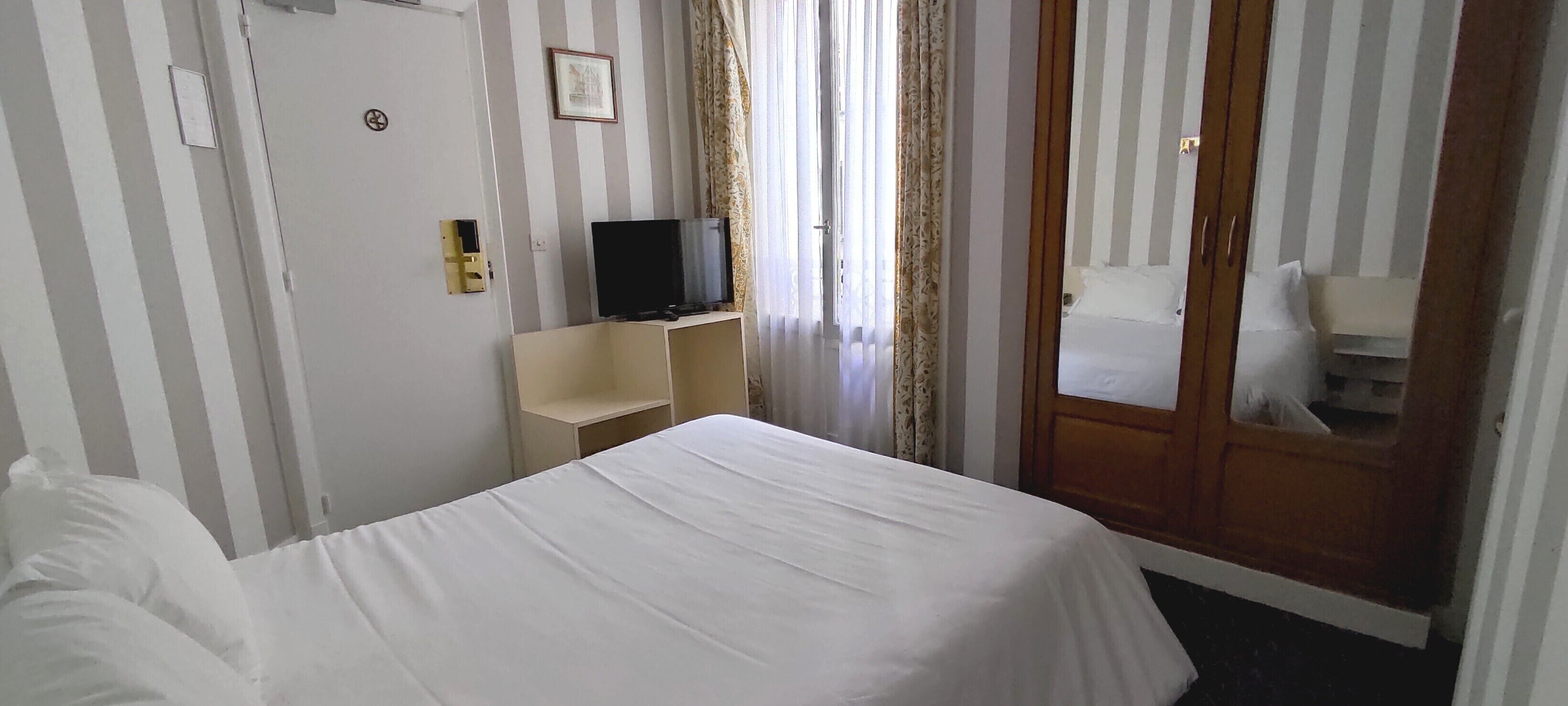A hotel room with a bed, a television on a stand, a wooden wardrobe, and a door with a mirror.