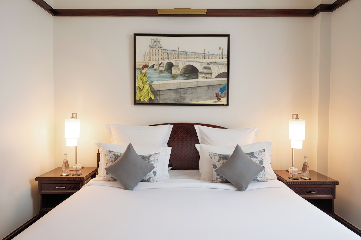 A hotel room with a large bed, two bedside tables, a painting of a bridge, and two lamps.