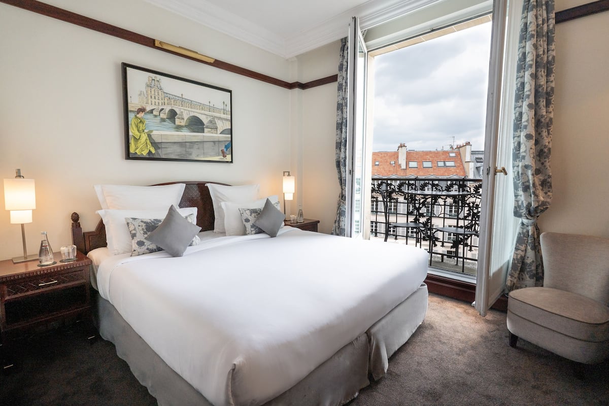 A hotel room with a large bed, two bedside tables, a chair, a balcony with a railing, and a painting on the wall.
