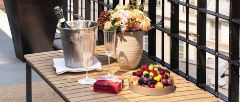Un balcón con una mesa lista para dos personas, que incluye una botella de champán, copas, un florero con flores y un frutero. Una planta en maceta está a la izquierda, y la Torre Eiffel es visible al fondo.