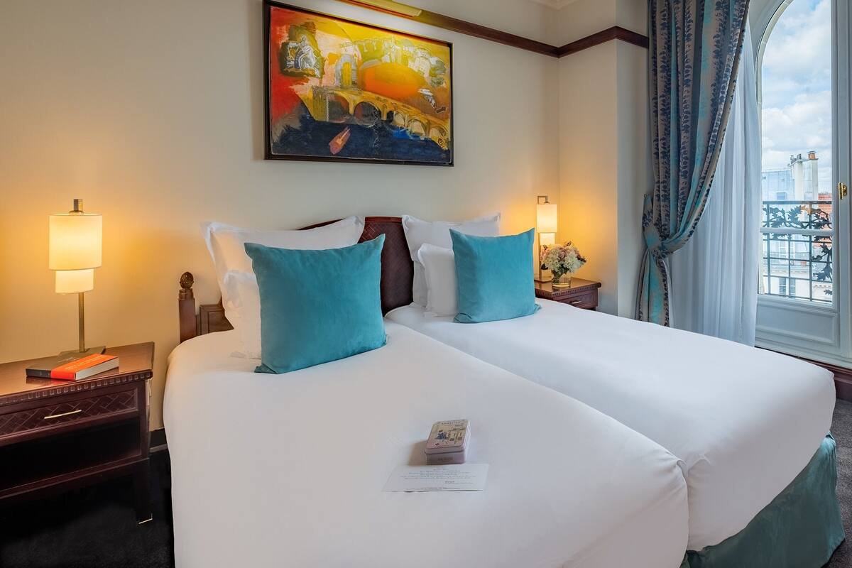 A hotel room with two beds, a painting on the wall, a window with curtains, and a nightstand with a lamp and a book.