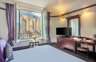 Una habitación de hotel con un ventanal grande, una cama, un escritorio con computadora, un televisor y una mesita con un jarrón de flores.
