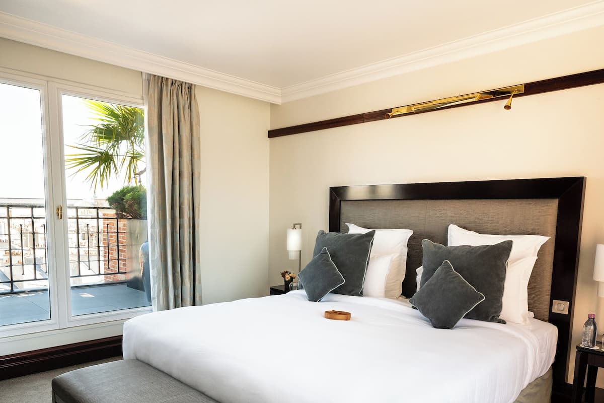 A hotel room with a large bed, a bench, a nightstand, a lamp, and a view of the outdoors.