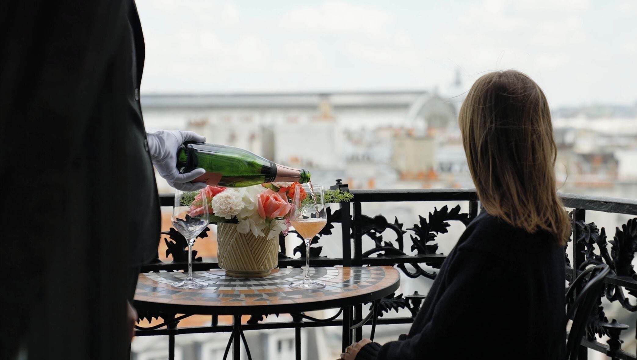 A person pouring champagne into a glass on a balcony with a table set with flowers and a vase.