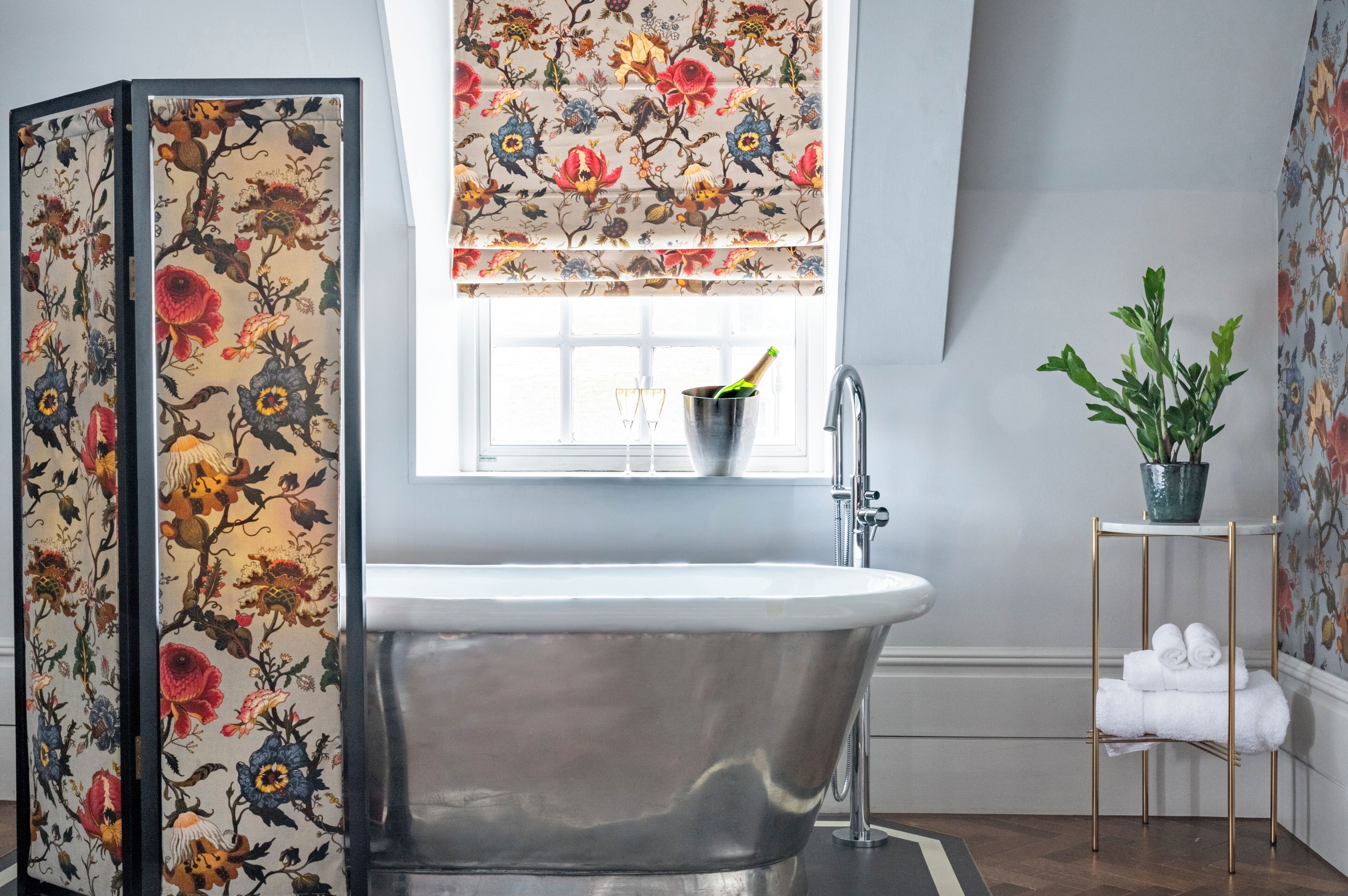 Une salle de bain avec une baignoire sur pieds, une fenêtre avec un store à motifs floraux, une plante en pot et un porte-serviettes.