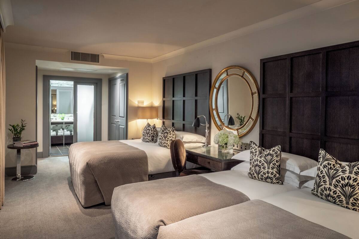 A hotel room with two beds, a large mirror, a desk with a lamp, and a view of the bathroom.