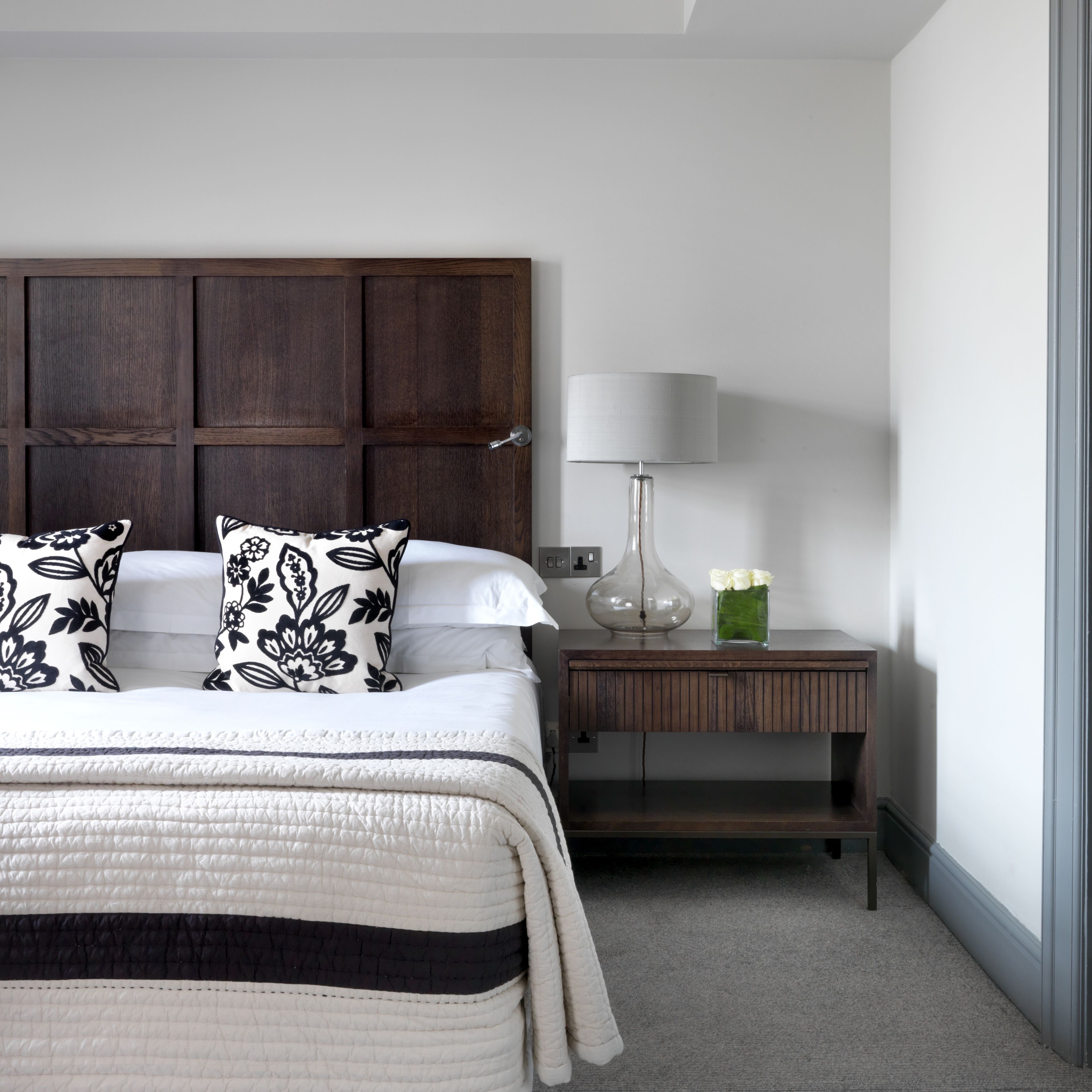 A bedroom with a large bed, a nightstand with a lamp, and a vase with flowers.