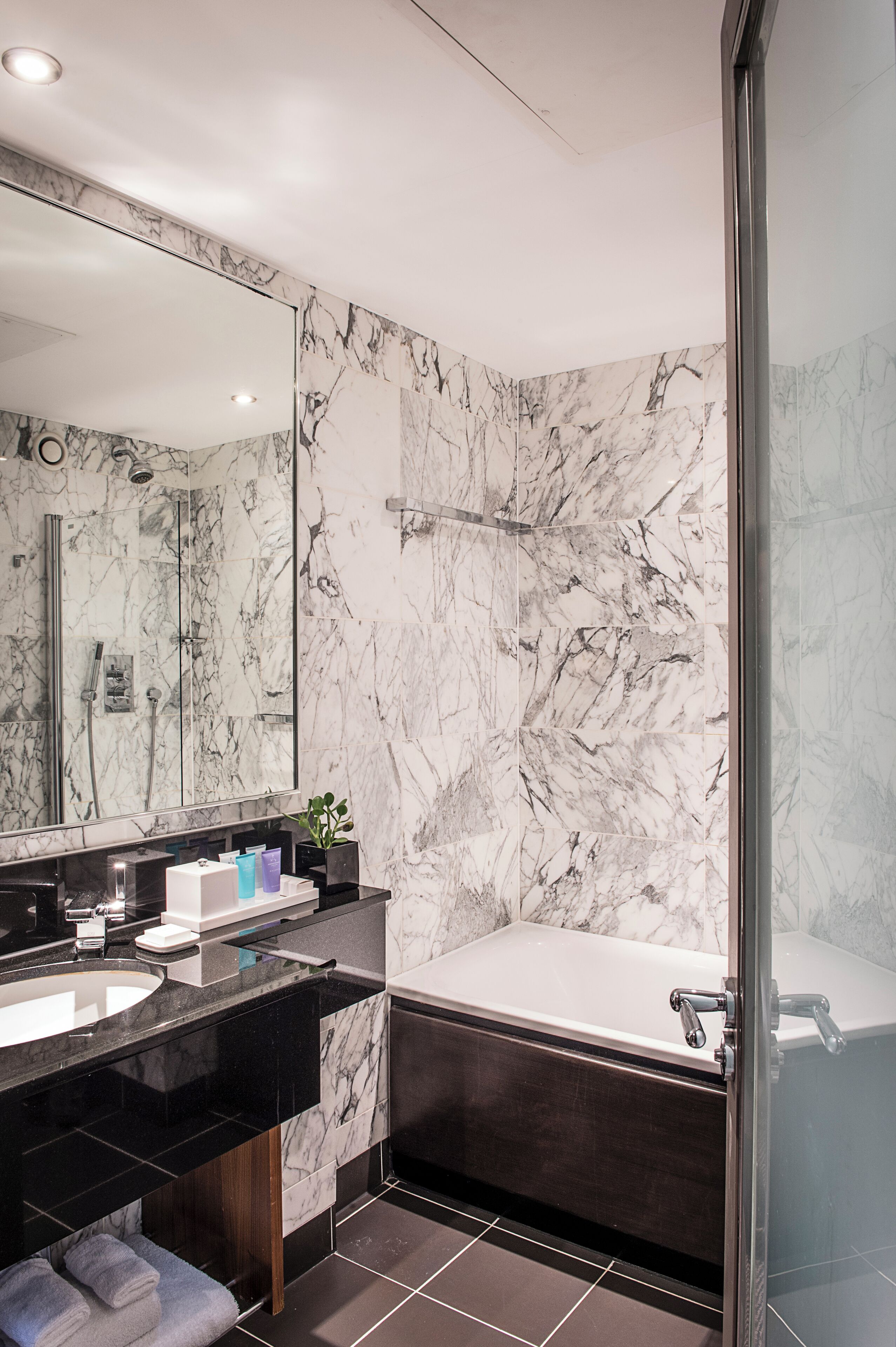 A modern bathroom with a marble wall, a large mirror, a black vanity, a bathtub, and a glass shower enclosure.