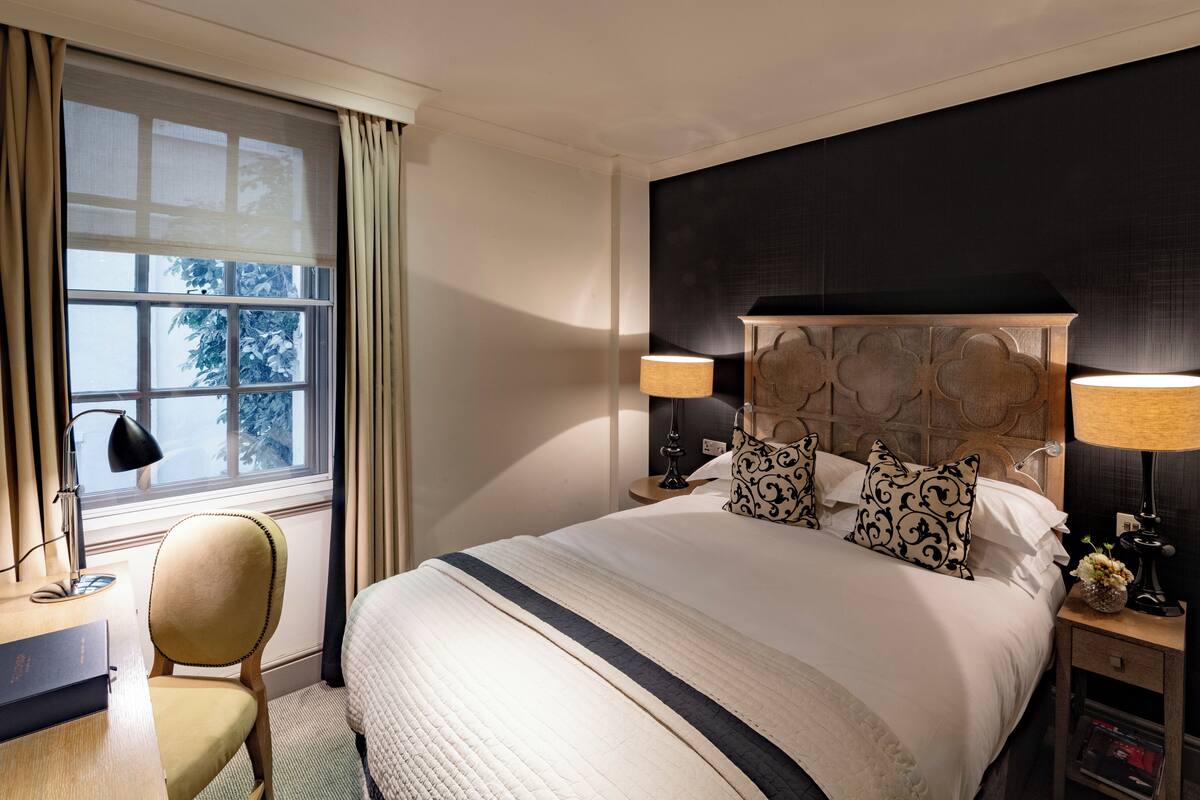 A hotel room with a large bed, two bedside lamps, a desk with a chair, and a window with curtains.
