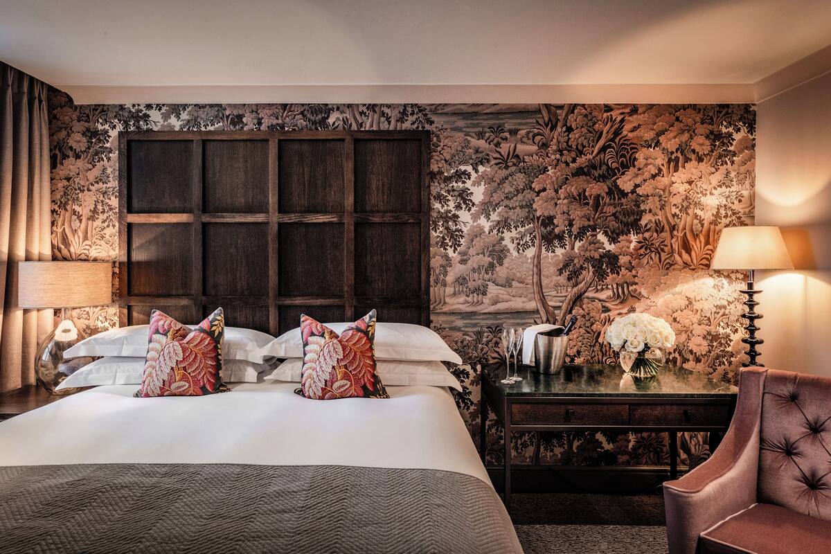 A hotel room with a large bed, two bedside tables with lamps, a chair, and a floral wallpaper.