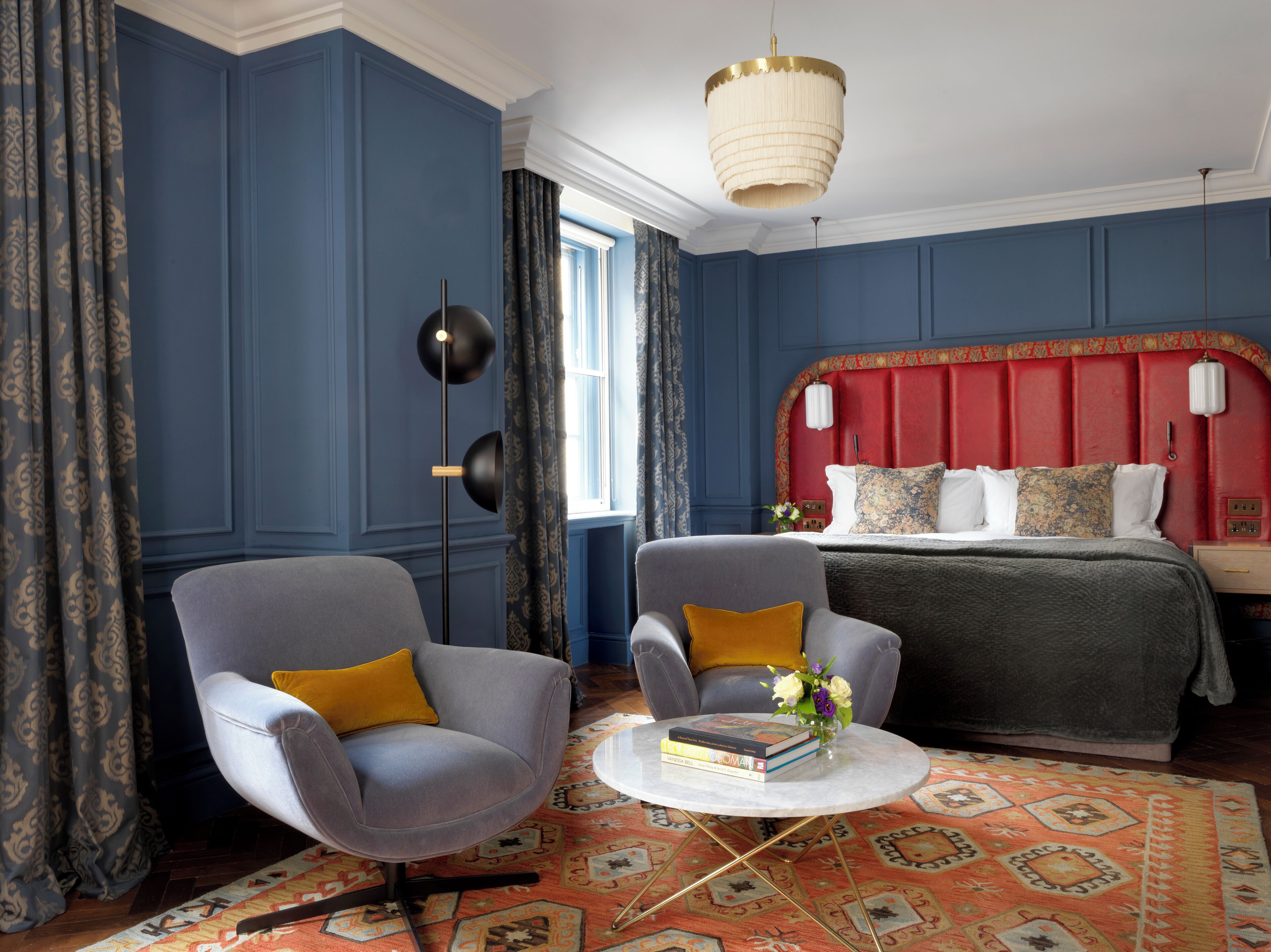 A bedroom with a red headboard, two grey armchairs, a round coffee table with books, and a patterned rug.