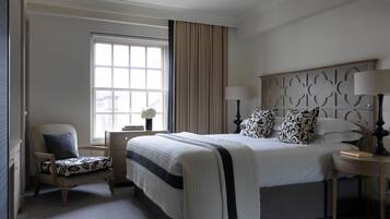 Ein ordentlich eingerichtetes Hotelzimmer mit einem großen Bett, einem Sessel, einem Nachttisch und einem Fenster mit Vorhängen.