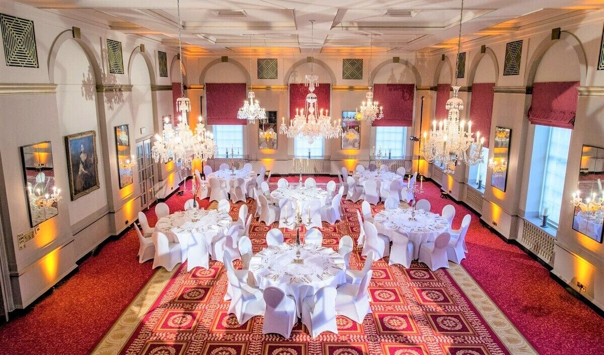 A banquet hall with round tables set for a formal event, featuring chandeliers and red carpet flooring.