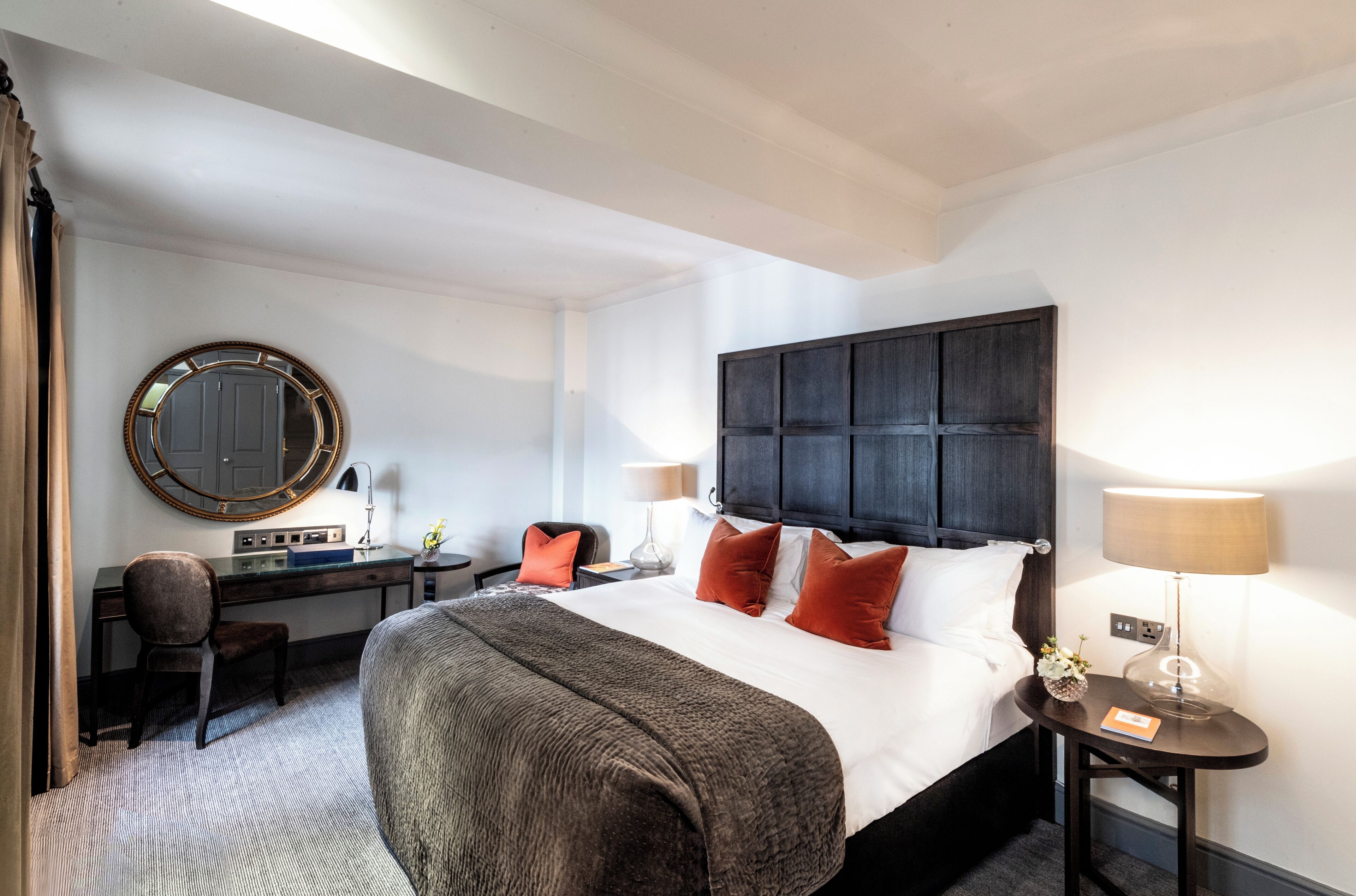 Een hotelkamer met een groot bed, een bureau met een stoel, een ronde spiegel en een nachtkastje met een lamp.