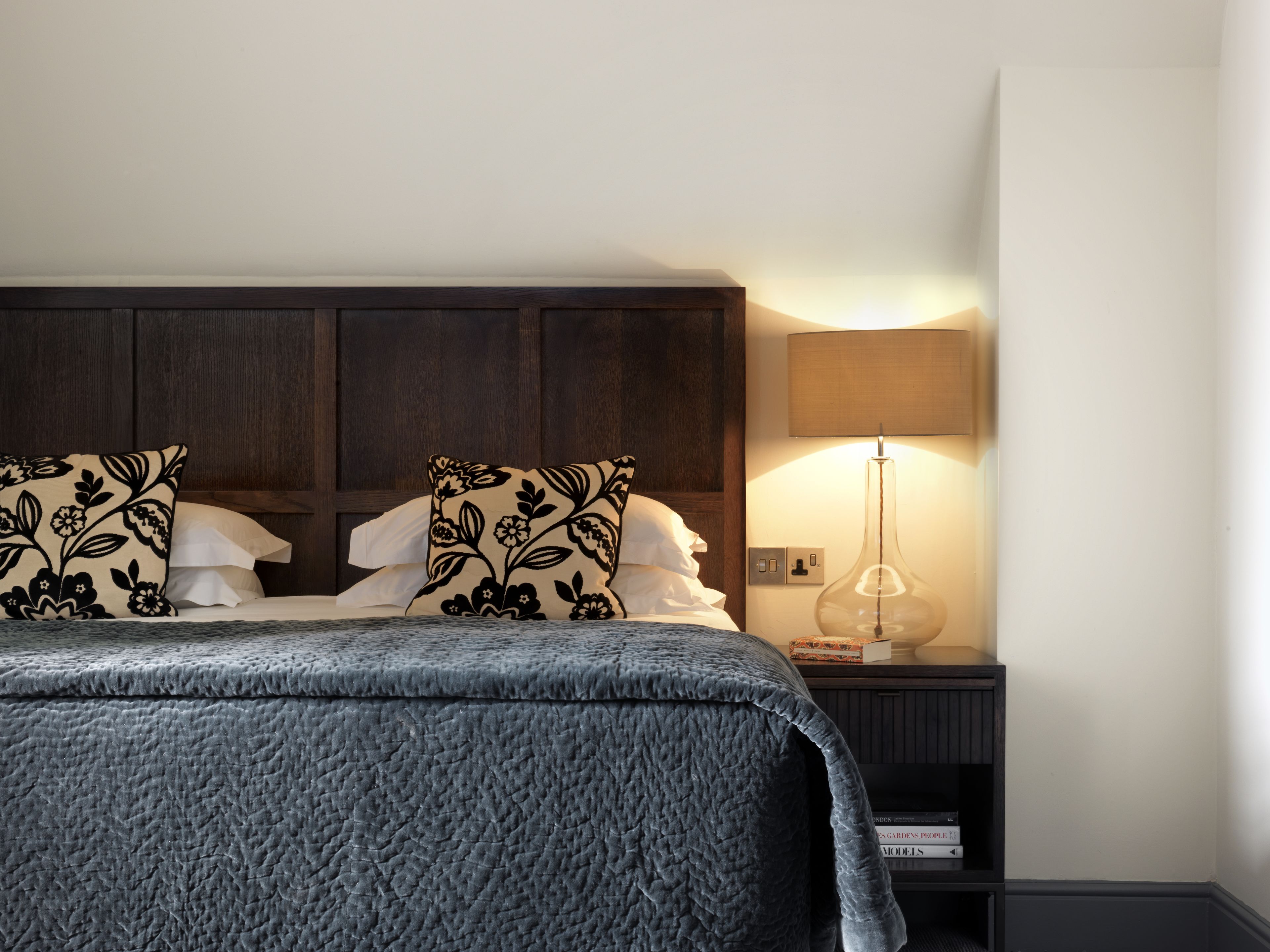 Ein ordentlich gemachtes Bett mit einer dunkelgrauen Decke, zwei dekorative Kissen mit einem schwarzen Blumenmuster, einer Nachttischlampe und einem kleinen Bücherregal mit Büchern.