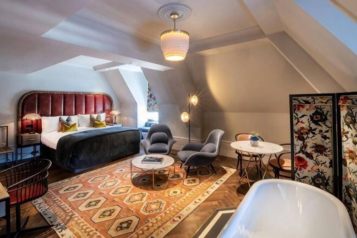 A modern loft bedroom with a sloped ceiling, a large bed, a seating area with a coffee table, and a view of the city.