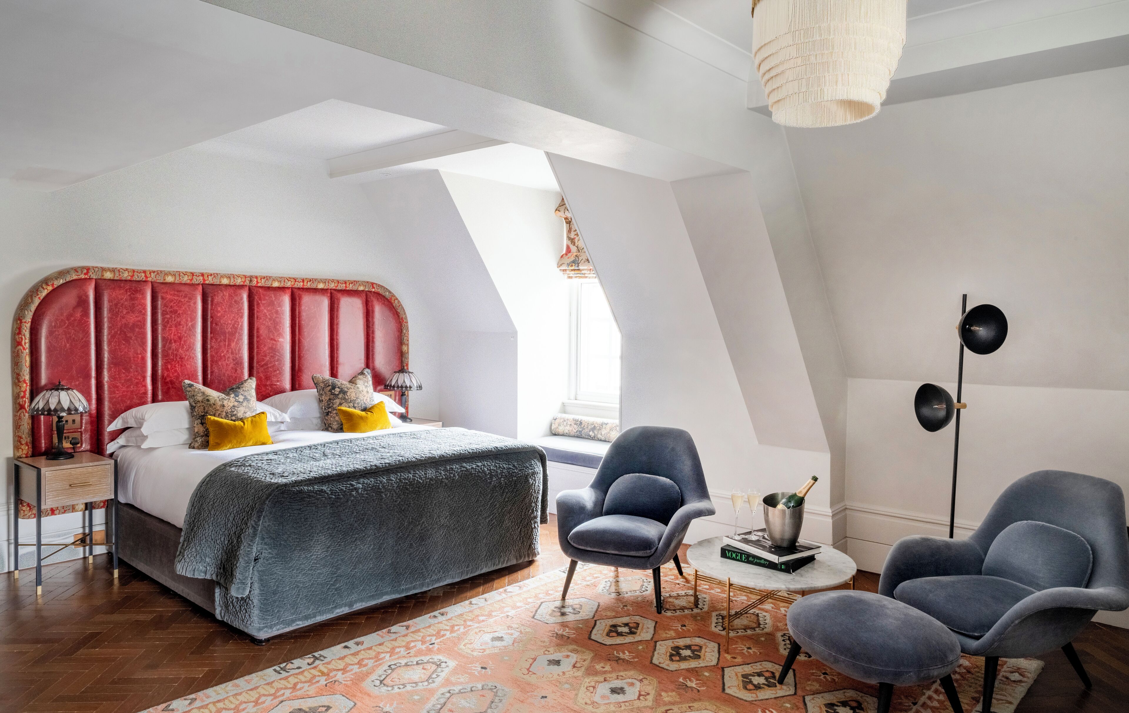 Ein modernes Schlafzimmer mit einem roten Kopfteil, einem großen Bett, zwei Sessel, einem kleinen Tisch mit einem Buch und Champagner sowie einer Stehlampe.