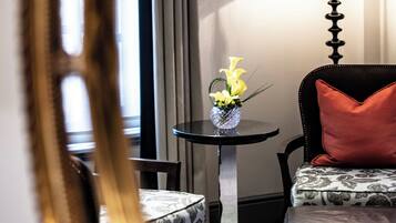 A hotel room with a round table, a chair, a sofa with a patterned cushion, a lamp, and a vase with flowers.