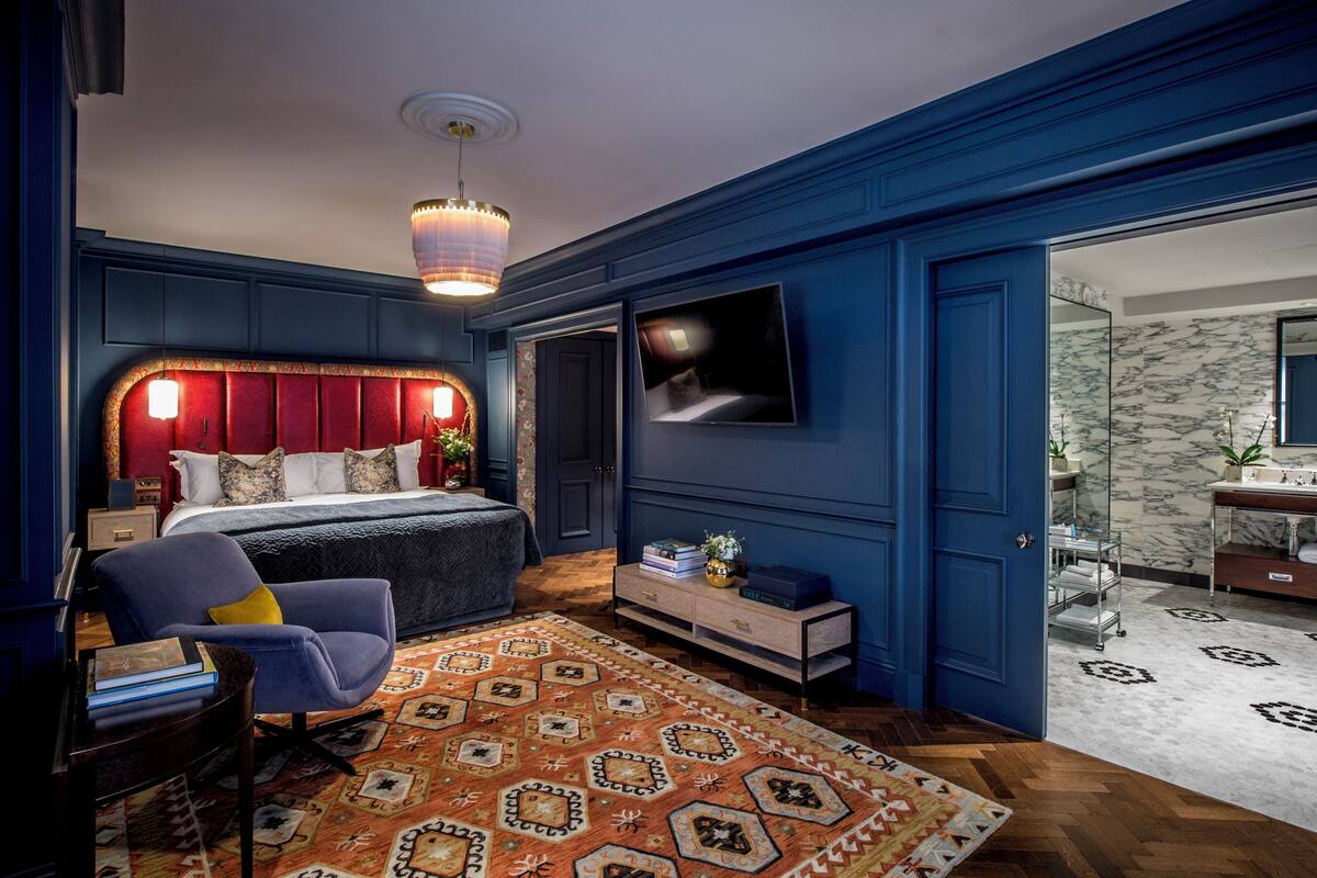 A bedroom with a bed, a chair, a TV, and a bathroom visible through an open door.