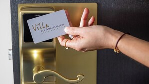 A hand holding a card with "Villa Des Ambassadeurs HOTEL PARIS" written on it, in front of a golden door handle. - Villa des Ambassadeurs (Paris)