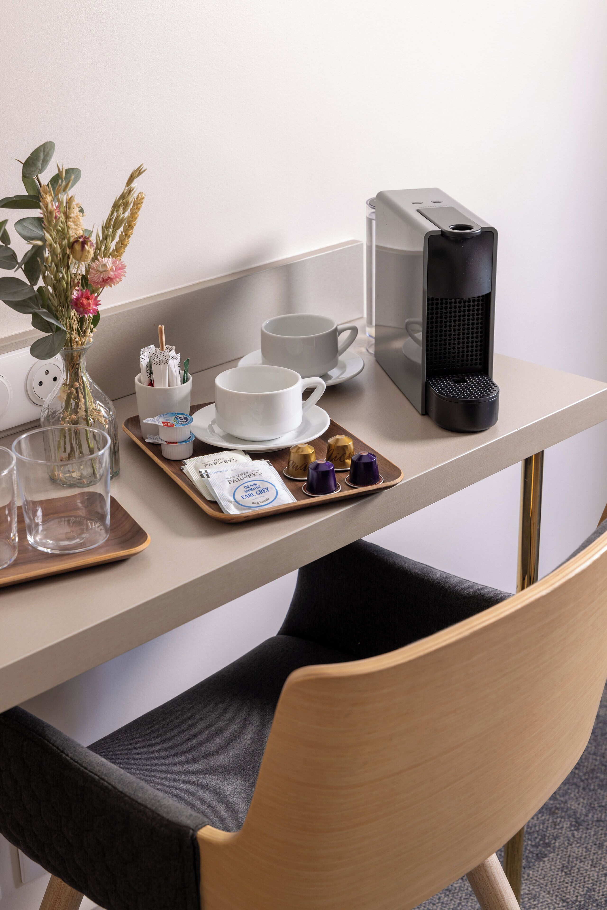 Una habitación de hotel con kitchenette, un comedor con una mesa dispuesta para dos personas, cafetera y un florero con flores.