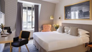 A hotel room with a large bed, a desk with a chair, a window with curtains, a framed picture on the wall, and a lamp. - Villa des Ambassadeurs (Paris)