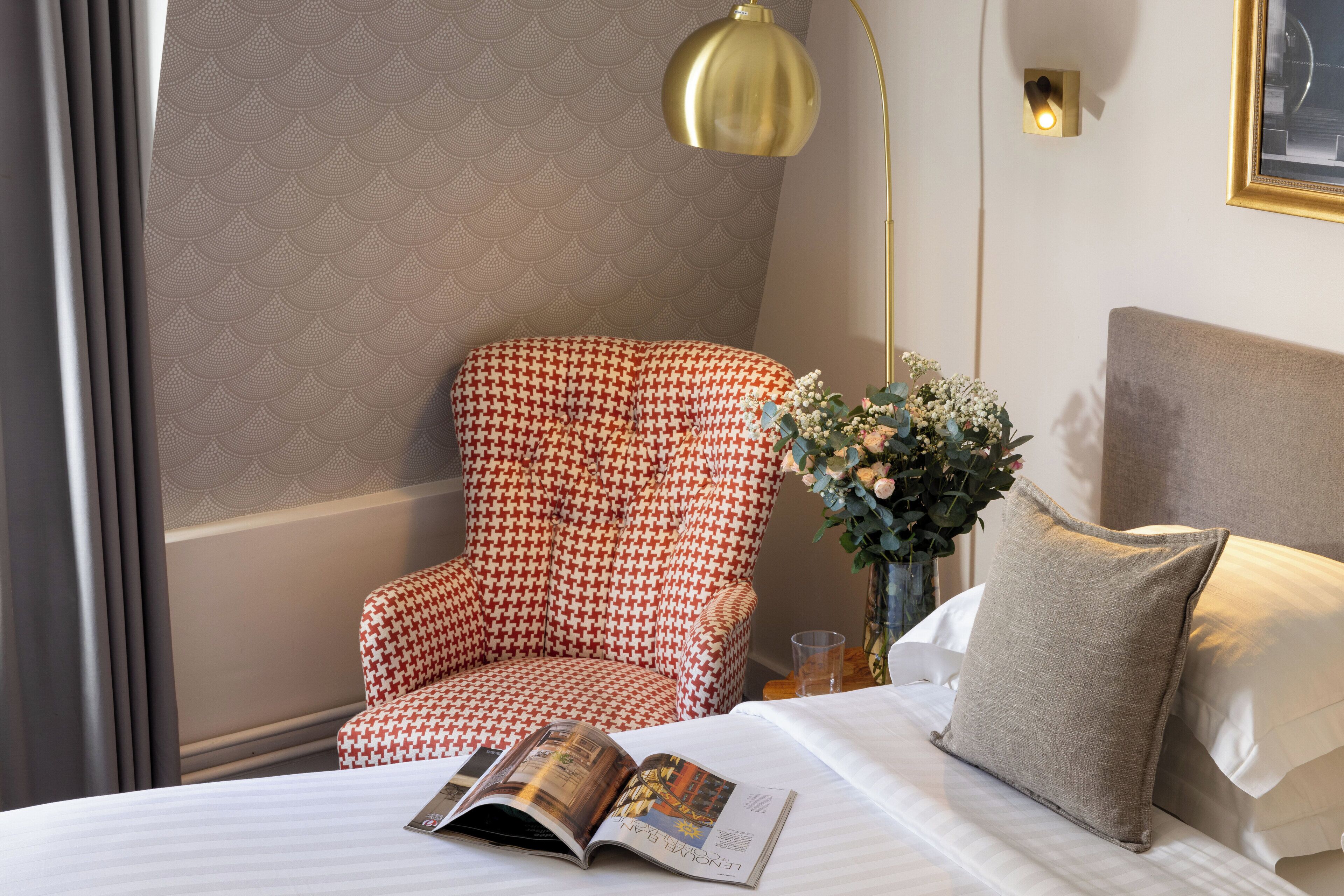 Une chambre d’hôtel avec un fauteuil à motifs, une table de chevet ornée d’un vase de fleurs et un magazine posé sur le lit.
