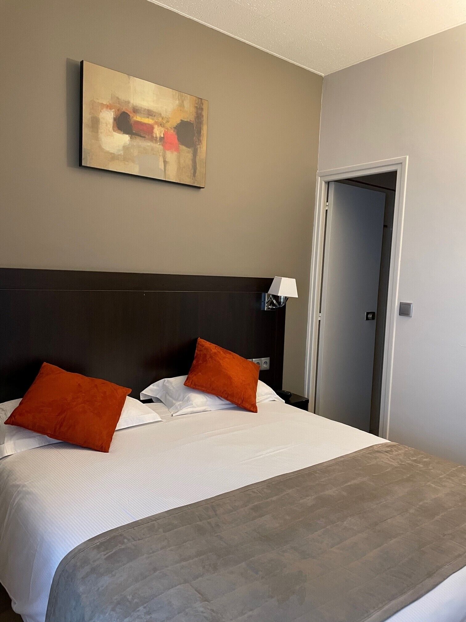 Habitación de hotel con una cama grande, dos almohadas anaranjadas, una colcha blanca, un plaid gris, un cuadro abstracto colgado en la pared, una lámpara de noche y una puerta.