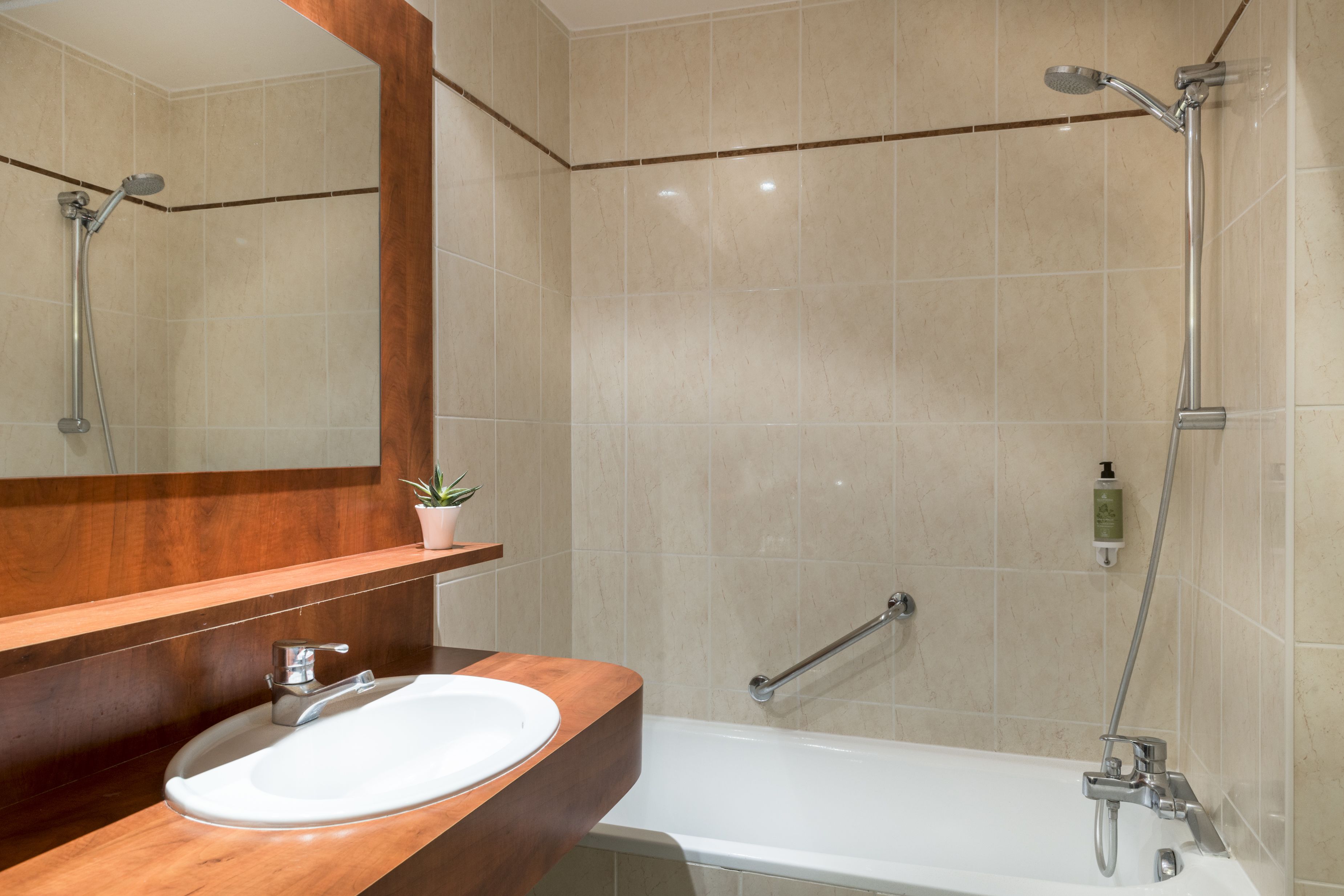 A bathroom with a wooden vanity, a white sink, a mirror, a shower with a handheld showerhead, and a handheld showerhead mounted on the wall.