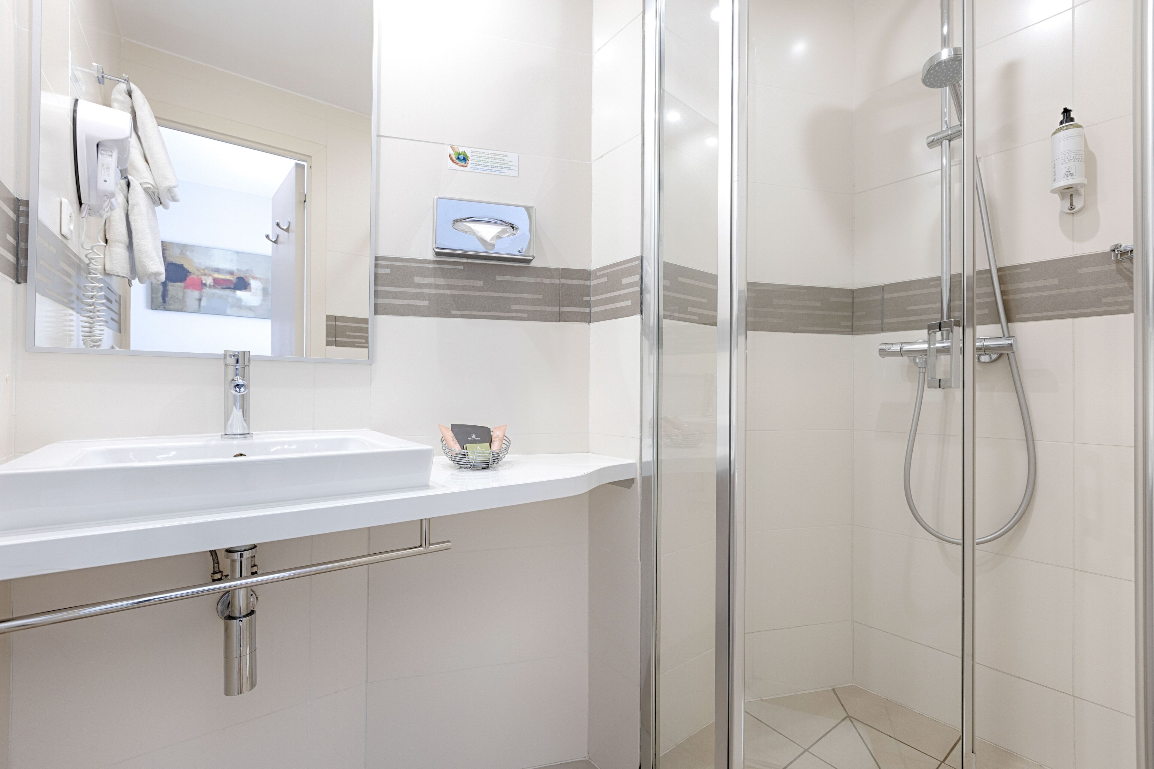 Un baño moderno con lavamanos blanco, un espejo, toallero, ducha con puerta de vidrio y dispensador de jabón.
