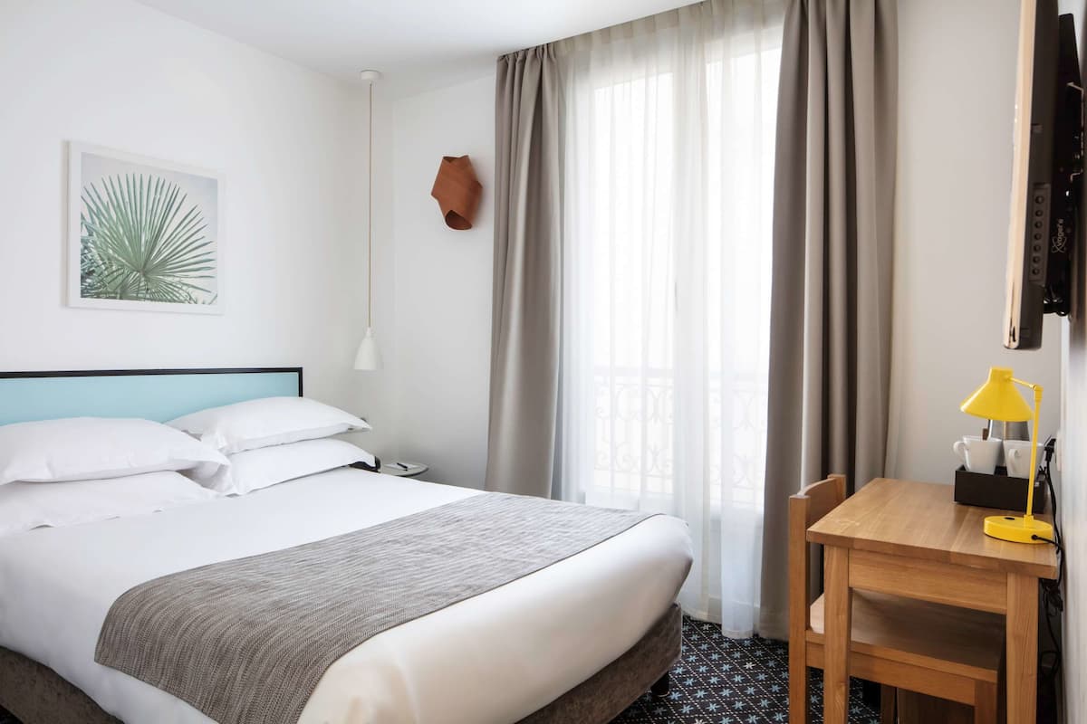 A hotel room with a bed, a desk, a lamp, a wall-mounted TV, and a window with curtains.